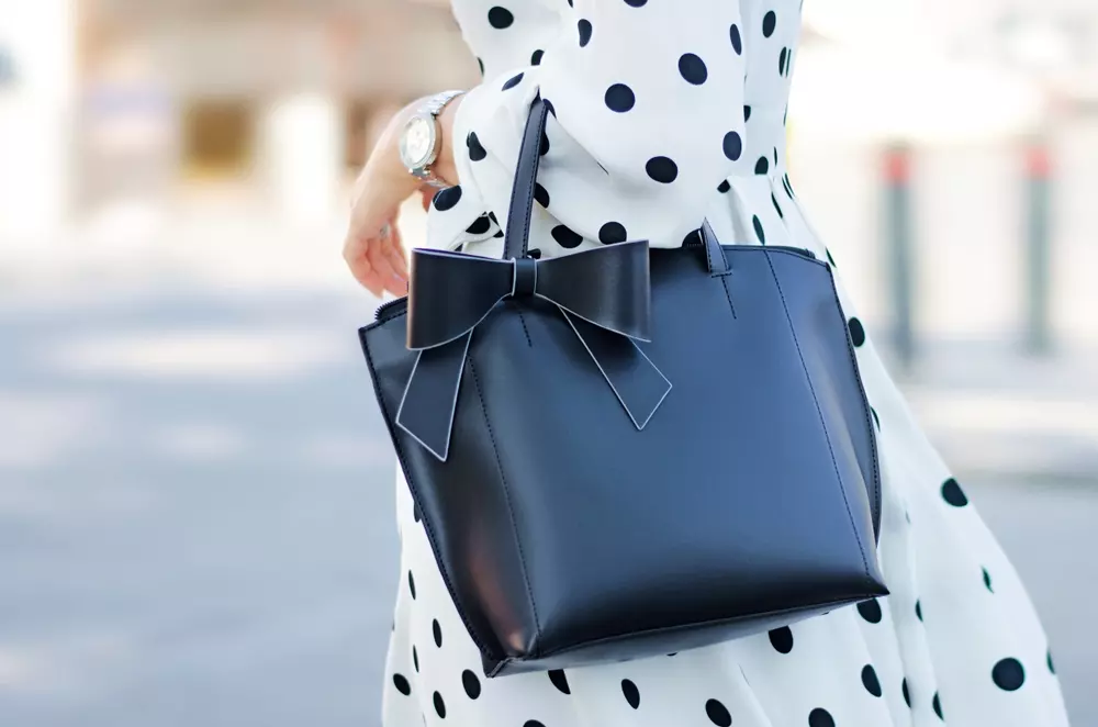 Bolsa preta feminina com laço decorativo usado por uma mulher de vestido branco com bolinhas pretas, em ambiente urbano ao ar livre.