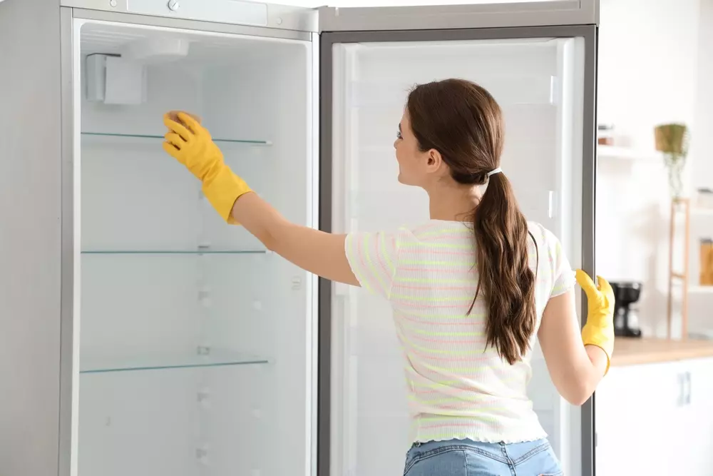 Mulher limpando interior de geladeira com luvas amarelas na cozinha moderna, cuidando da limpeza doméstica. Enfoque em higiene e manutenção de eletrodomésticos.