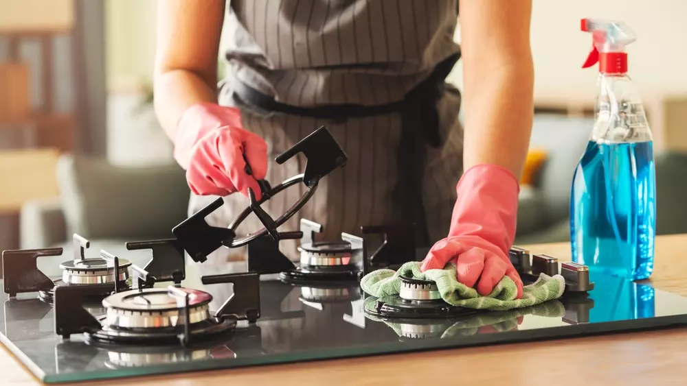Pessoa limpando fogão na cozinha como parte da limpeza da cozinha, usando luvas de borracha e paninho. Ambiente doméstico limpo e organizado.