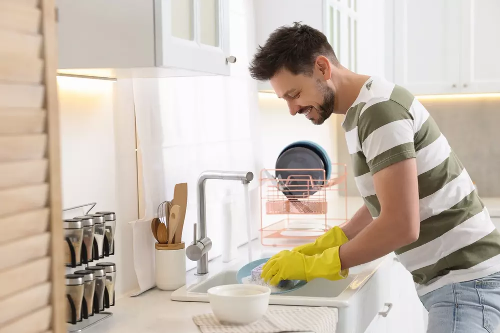 Homem feliz lavando louça na cozinha, que é parte da limpeza da cozinha, com utensílios e louça limpa ao fundo.