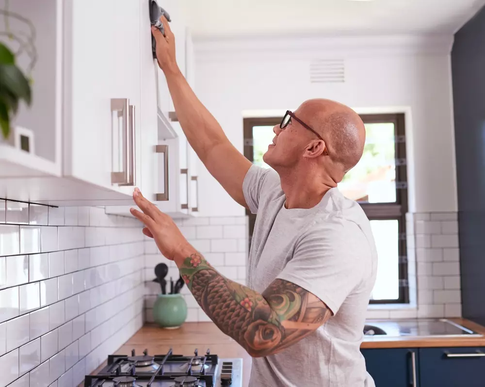 Homem limpando armário de cozinha moderno com armário branco e bancada de madeira, cozinha bem iluminada.