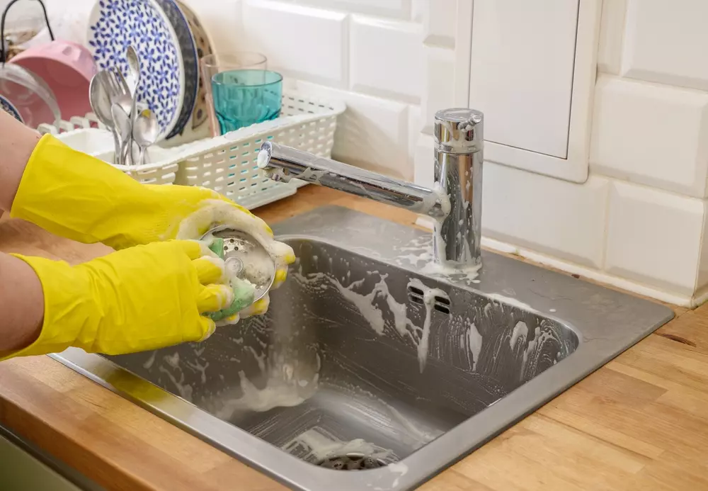 Pessoa limpando a pia da cozinha com luvas amarelas e produtos de limpeza, promovendo a limpeza da cozinha e do ralo.