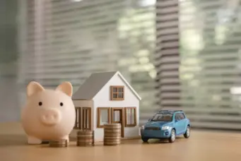 Imagem ilustrando o planejamento financeiro em um contexto de Selic alta para compra do primeiro imóvel, com porquinho, casa de brinquedo, pilhas de moedas e carro em uma mesa.