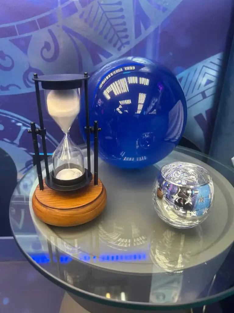 Detalhes do Quarto Eternidade do BBB 26 com uma ampulheta, uma bola de vidro azul e um vaso de vidro na decoração ambiente moderna.