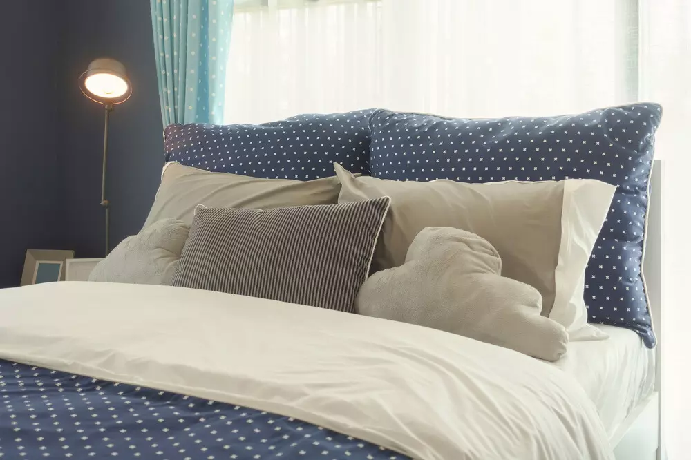 Cama de casal com roupa de cama bege e azul com estampa de bolinhas, decorada com várias almofadas confortáveis, em um quarto bem iluminado.