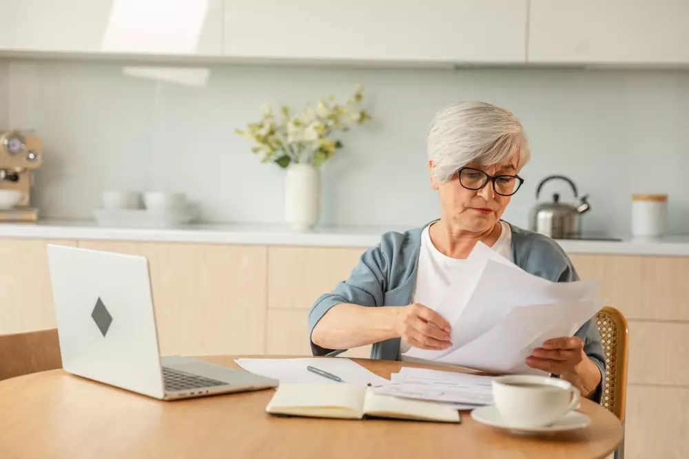 Mulher idosa analisando documentos de IPTU pago em duplicidade, sentado à mesa na cozinha com laptop, caderno e café, preocupado com fatura em dupla.