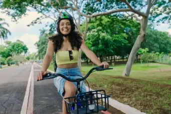 Jovem mulher sorridente pedalando de bicicleta em parque com árvores verdes e céu azul