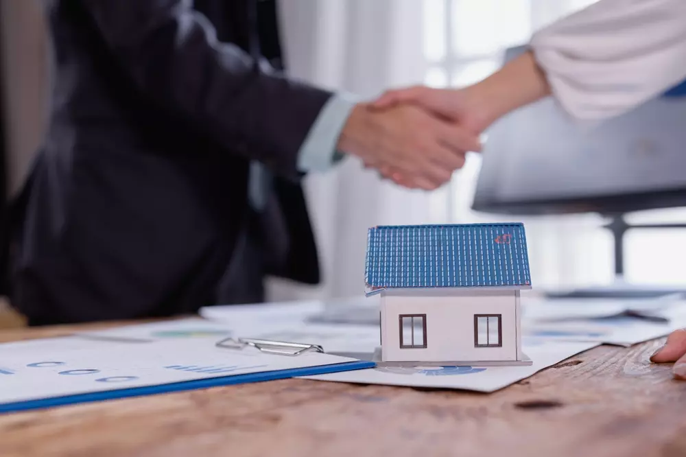 Acordo de compra e venda de imóvel com duas pessoas apertando as mãos, ao lado de uma maquete de casa sobre a mesa, simbolizando negociação imobiliária.