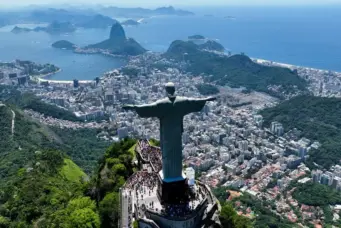 Vista aérea do Rio de Janeiro com o Cristo Redentor em destaque, mostrando bairros, florestas e a orla da cidade brasileira.