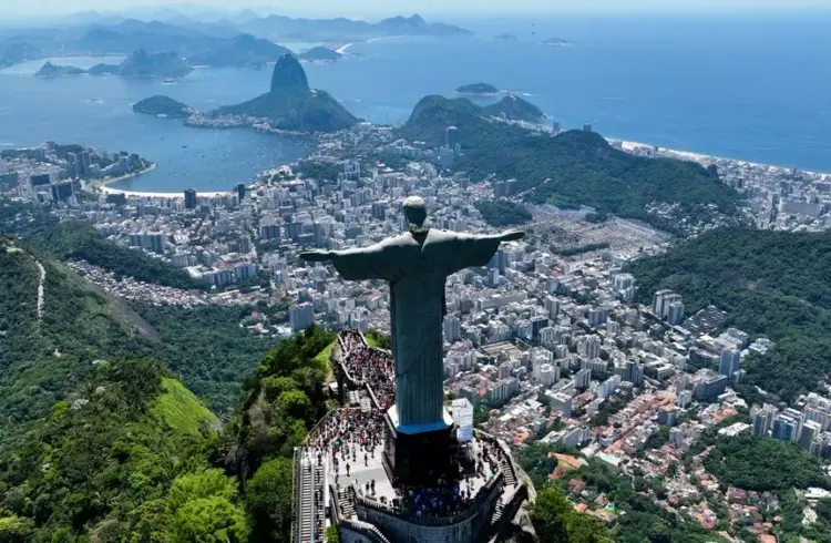 Vista aérea do Rio de Janeiro com o Cristo Redentor em destaque, mostrando bairros, florestas e a orla da cidade brasileira.