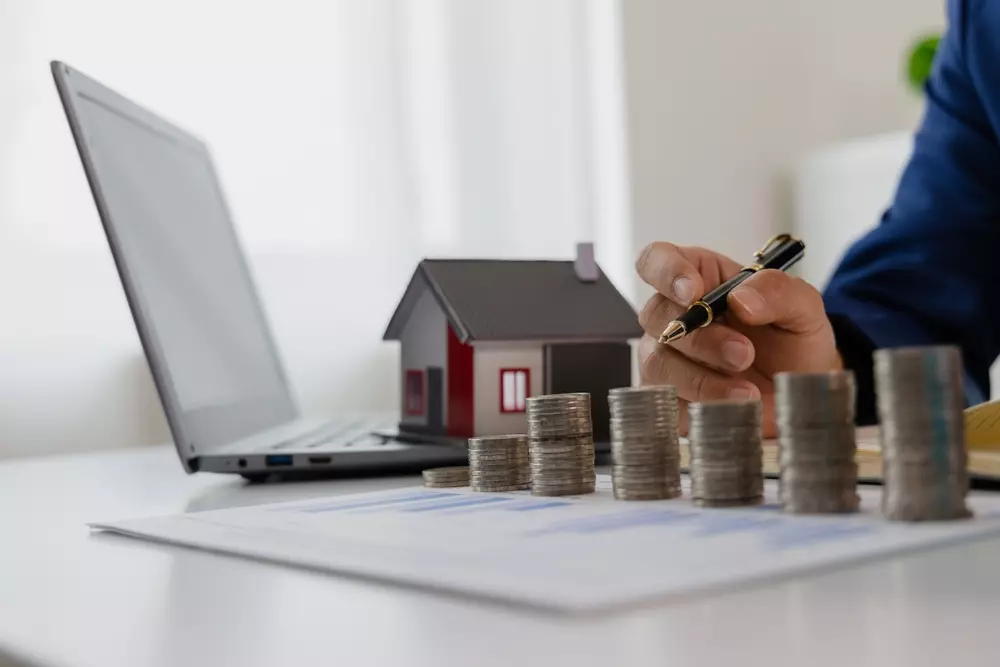 Pessoa fazendo planejamento financeiro para financiamento conjunto de imóvel, com blocos de moedas, computador e miniatura de casa, trazendo insights sobre capacidade de pagamento no longo prazo.