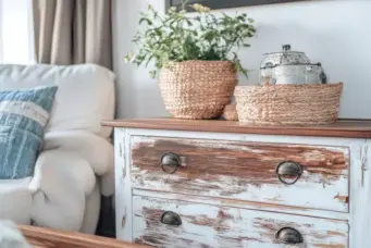 Decoração de ambiente com estilo shabby chic, destacando móvel de madeira com pintura desgastada, vasos de fibra natural com plantas e objetos vintage. Ideal para quem busca charme de casas de campo europeias.