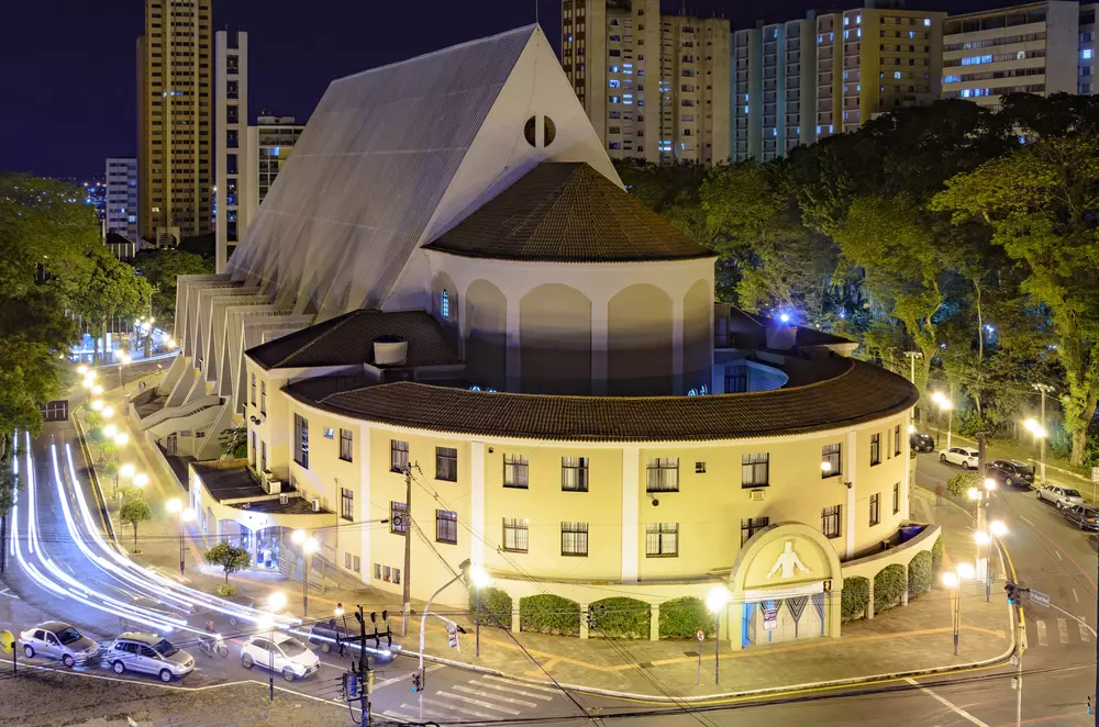 Catedral Metropolitana de Londrina iluminada à noite, com destaque para sua arquitetura moderna e telhado triangular, rodeada por árvores e veículos na cidade.