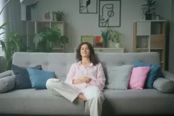 Mulher relaxada em sala de estar confortável com muitas plantas e decoração moderna, ilustrando os 5 tipos de riqueza na vida, como saúde, tempo, relacionamentos, conhecimento e bens materiais.