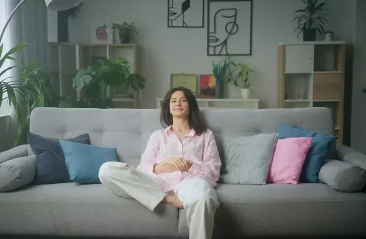 Mulher relaxada em sala de estar confortável com muitas plantas e decoração moderna, ilustrando os 5 tipos de riqueza na vida, como saúde, tempo, relacionamentos, conhecimento e bens materiais.