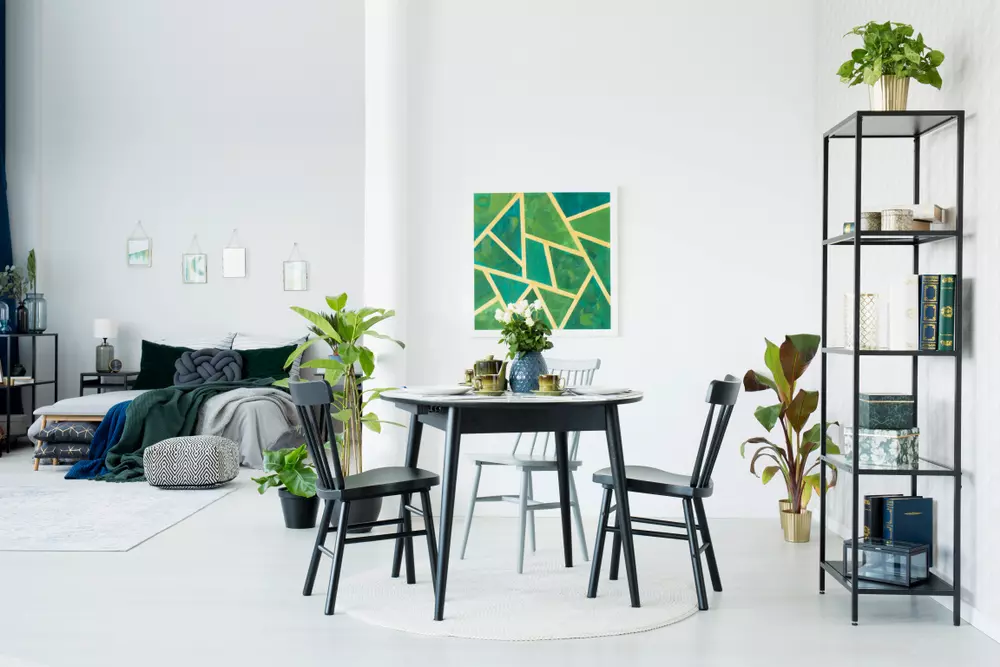 Sala de jantar moderna com mesa de jantar preta, cadeiras pretas, prateleira de metal, plantas decorativas, e parede branca com arte abstrata