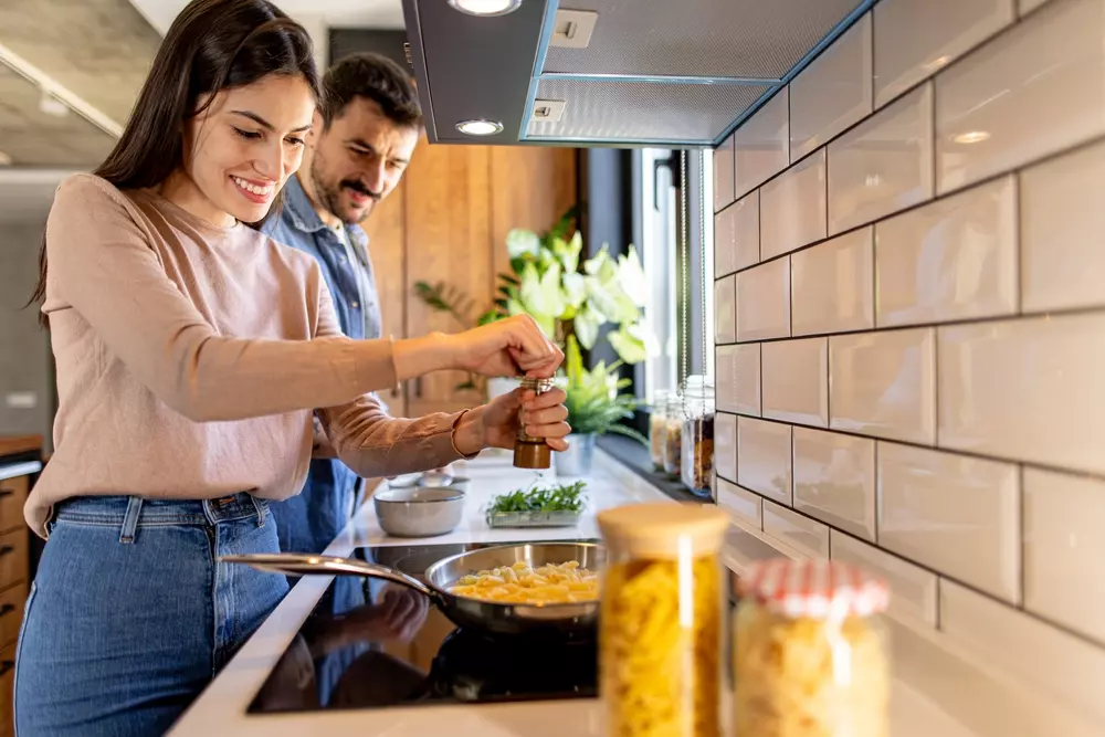 Casal preparando uma refeição na cozinha moderna com foco em um cooktop à gás, com animais decorativos no fundo, ambiente aconchegante e bem iluminado.