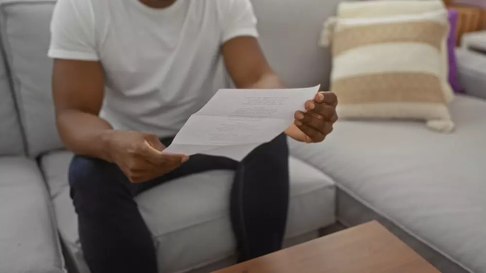 Pessoa lendo documento ou carta em ambiente doméstico, sentado em um sofá cinza com almofadas coloridas ao fundo.