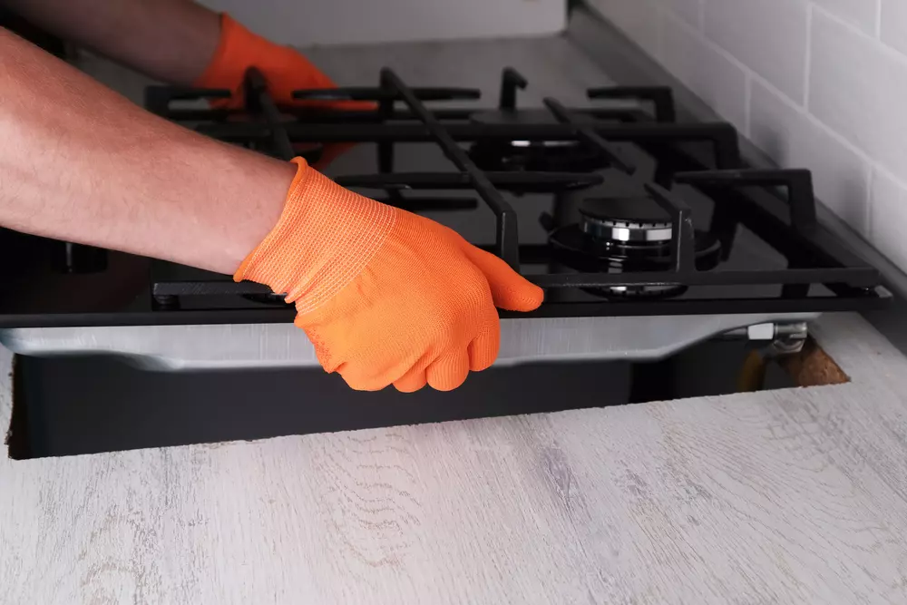 Pessoa instalando ou reparando cooktop de gás na cozinha com o uso de luvas laranja, em ambiente de cozinha com bancada de mármore claro e parede de azulejo branco.