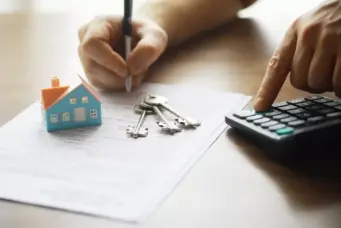 Pessoa assinando documentação relacionada à Faixa 4 ao lado de chaves e uma casa em miniatura, simbolizando processos imobiliários.