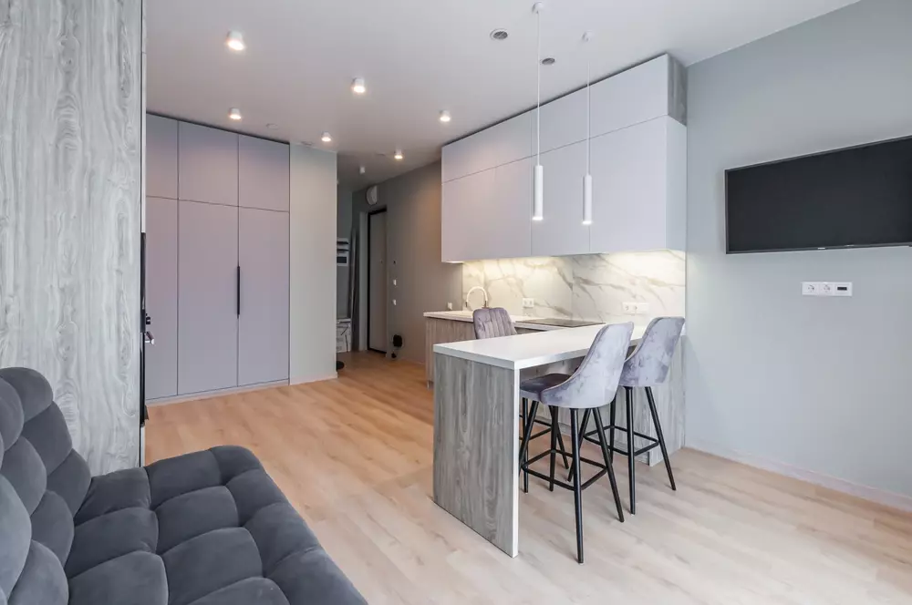 Uma sala moderna com móveis coloridos, incluindo um sofá cinza, cozinha compacta com bancada branca e cadeiras cinza, parede com TV e iluminação embutida.