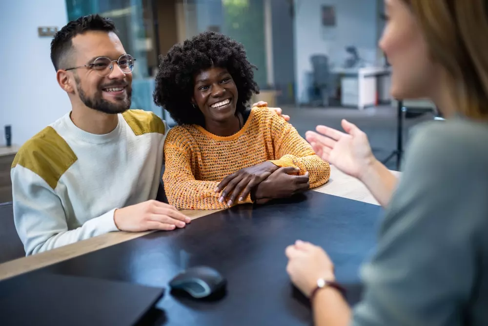 Três pessoas em uma reunião de negócios, incluindo uma mulher negra sorridente e um homem com barba e óculos, conversando com uma colega na frente deles em ambiente de escritório moderno