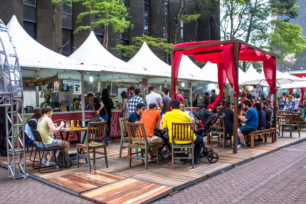 pessoas aproveitando feira de rua ao ar livre com mesas, tendas brancas e estrutura de madeira, na cidade, ambiente social e gastronômico ao ar livre