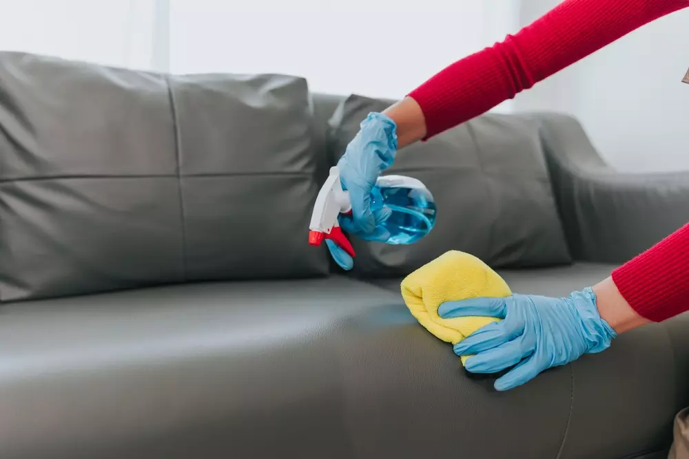 Pessoa limpando sofá de couro com pano amarelo e spray de limpeza, destacando o uso do couro na decoração de ambientes.