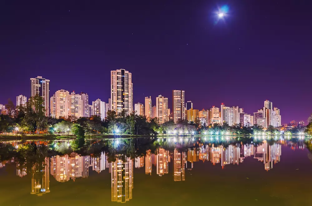 Vista noturna do centro de Londrina com arranha-céus iluminados refletidos no lago sob um céu estrelado, destacando a modernidade e beleza da cidade.