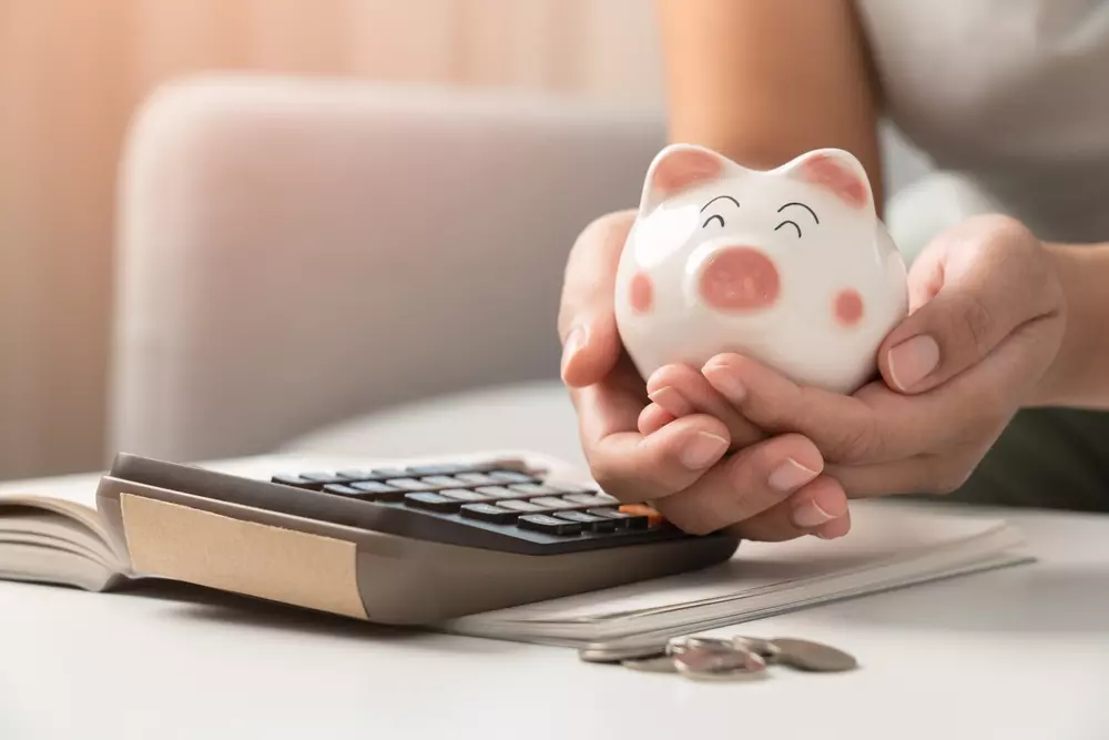 Pessoa segurando uma vaquinha de cerâmica com rosto sorridente, cartão de poupança e moedas ao lado, representando a reserva de emergência financeira.