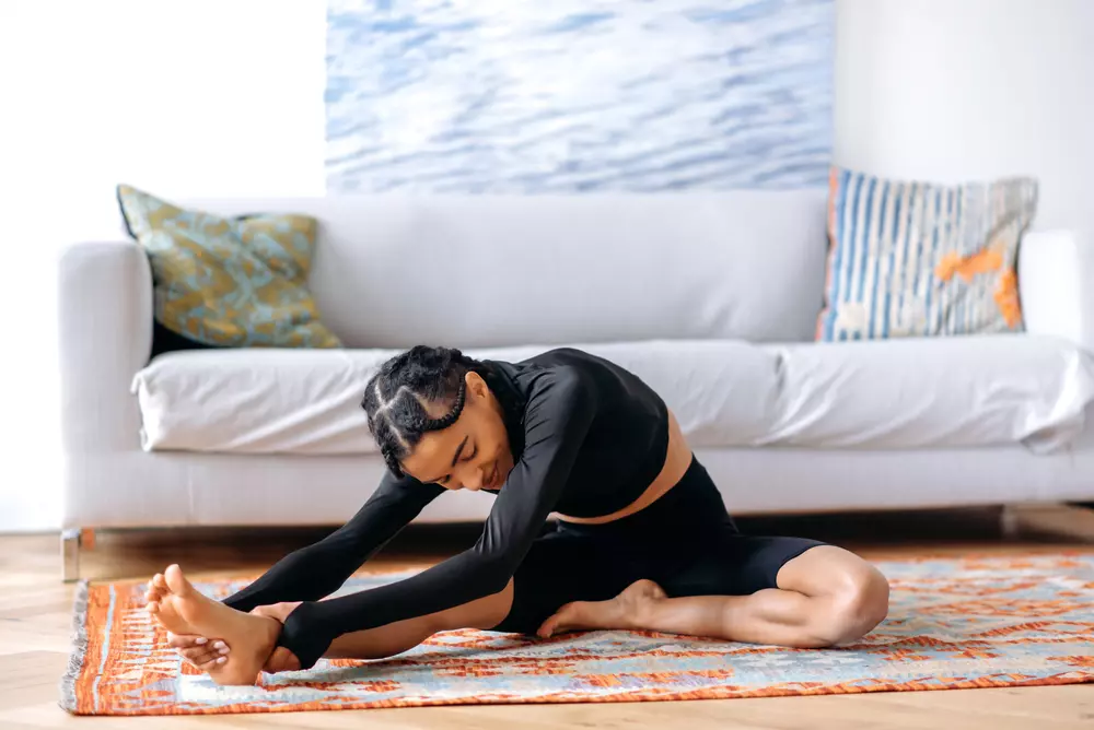 Mulher praticando yoga dentro de casa, demonstrando força física e flexibilidade, no ambiente moderno com sofá e decoração colorida ao fundo.