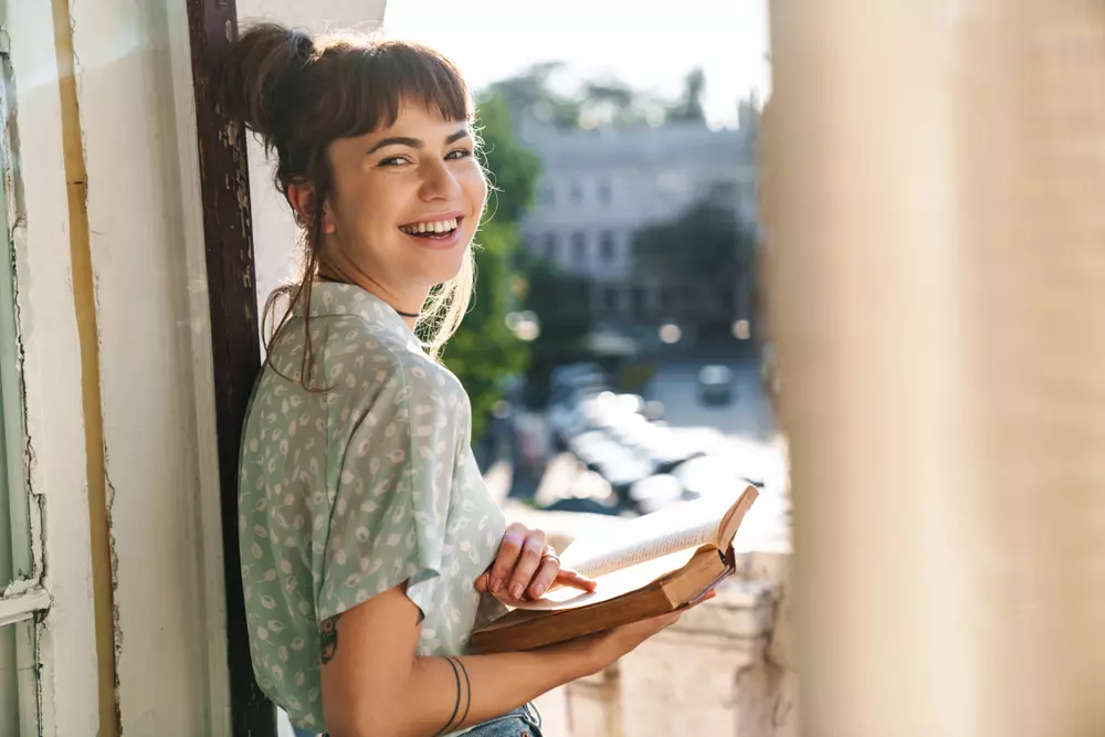 Mulher sorrindo segurando um livro, simbolizando a riqueza mental e o bem-estar emocional, destaque para o equilíbrio e clareza na vida.
