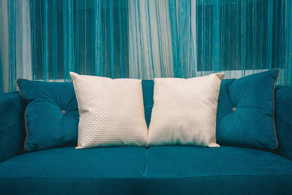 Um sofá azul decorado com almofadas brancas, localizado em uma sala moderna com parede de cortinas azuis e detalhes em destaque.