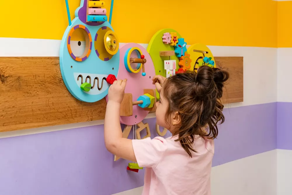 Criança interagindo com uma decoração educativa colorida e interativa em uma parede de escola ou sala de aula, estimulando o aprendizado e o desenvolvimento infantil.