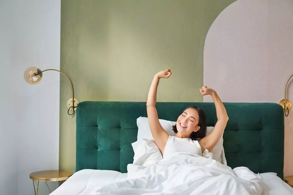 Mulher sorridente se alongando na cama em um quarto com paredes verdes e decoração moderna, ideal para escolher cores para o quarto.