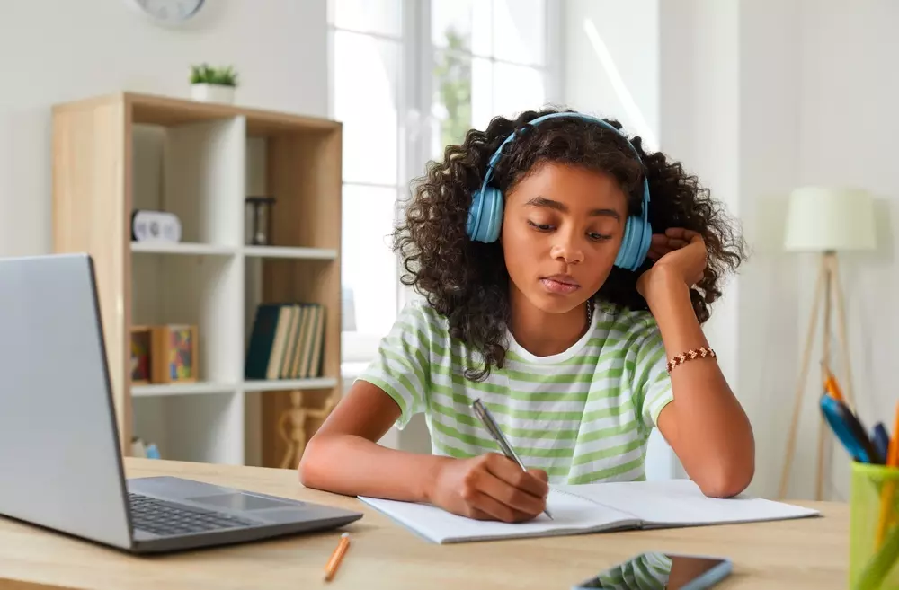 Jovem estudante de área de estudo assistindo aula online com fones de ouvido e fazendo anotações em um caderno, em ambiente bem iluminado.