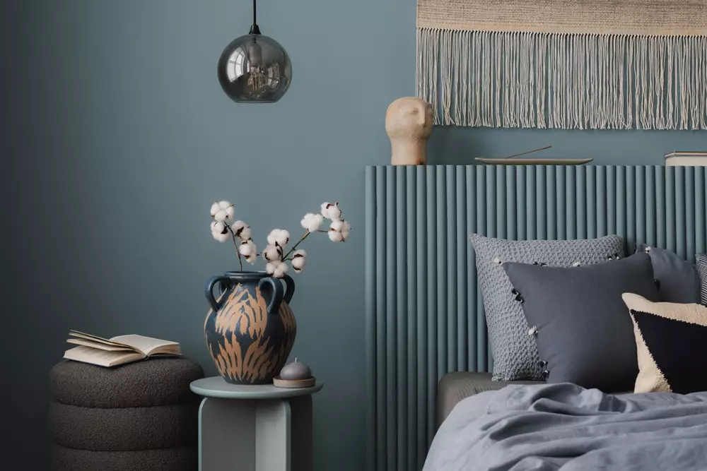Decoração de quarto com cores em tons de azul e cinza, incluindo parede azul, cabeceira azul, almofadas cinza e acessórios decorativos.