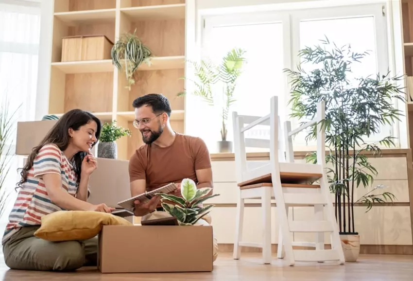 Casal alegre sentado no chão de uma sala bem iluminada, decorada com plantas e móveis de madeira, conversando e olhando fotos ou itens em um álbum.