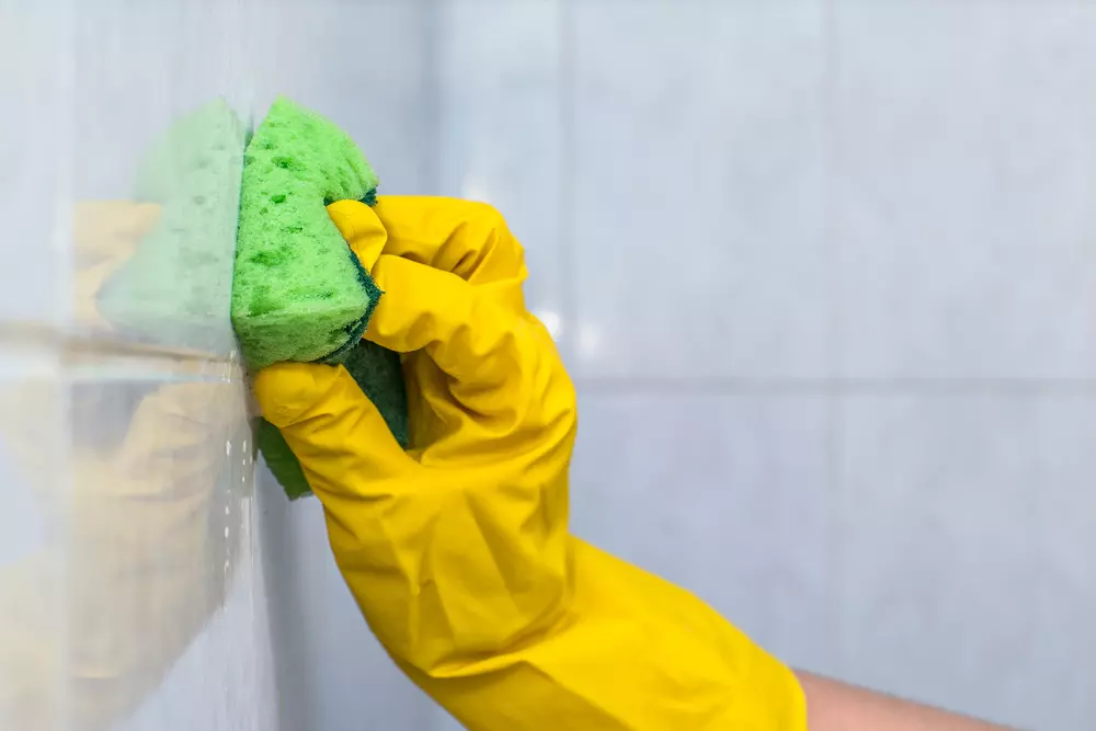 Mão de uma pessoa com luva de borracha amarela removendo resíduos difíceis na parede com esponja macia verde.