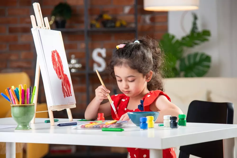 Menina pintando uma tela com tinta vermelha em um espaço criativo, decorado com itens educativos e coloridos que estimulam a aprendizagem e a criatividade infantil.