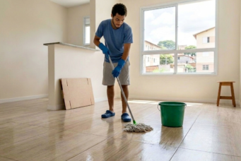 Homem realizando limpeza pós-obra em residência com piso de madeira, usando esfregão e balde, ambiente bem iluminado com janelas grandes.