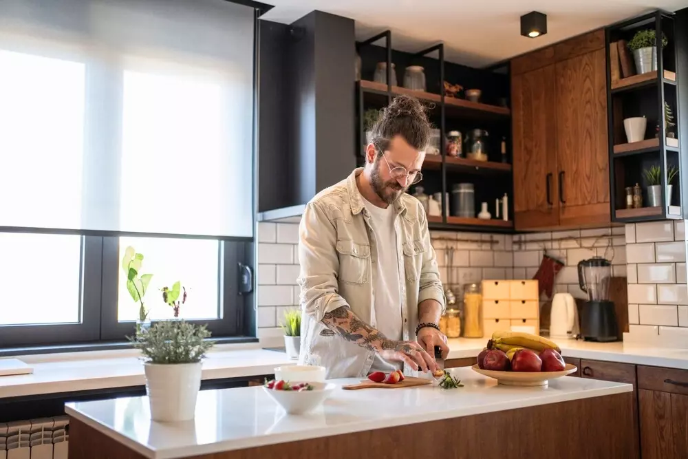 Homem preparando alimentos em uma ilha na cozinha moderna com armários de madeira, prateleiras abertas e janelas amplas para iluminação natural.