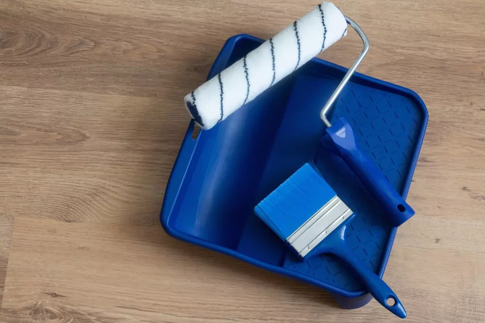 Itens de pintura, incluindo rolo, espátula e trincha, dispostos em uma bandeja azul sobre piso de madeira, ideal para reforma e decoração.