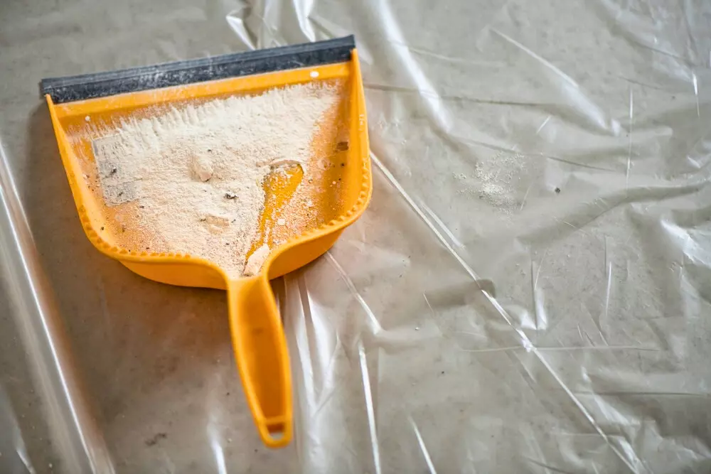 Limpeza pós-obra com pá de plástico amarela, pó de argamassa e plástico transparente na superfície, ideal para remover resíduos de construção.