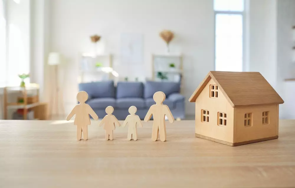 Casa de miniatura de madeira ao lado de figuras de pessoas também de madeira, simbolizando uma família, em um ambiente aconchegante e bem iluminado.