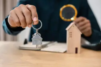 Pessoa segurando uma chave com uma casa de metal e uma casa de madeira ao fundo, simbolizando venda de imóveis na faixa 4 do Minha Casa Minha Vida.