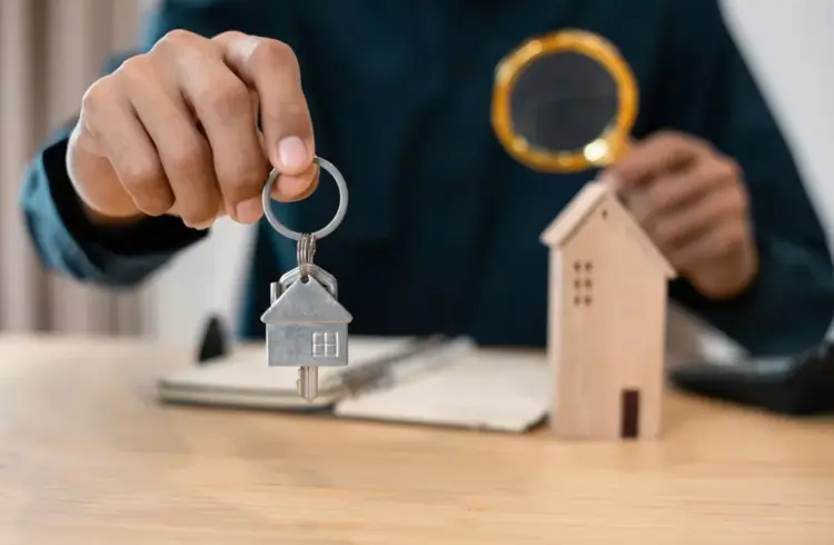 Pessoa segurando uma chave com uma casa de metal e uma casa de madeira ao fundo, simbolizando venda de imóveis na faixa 4 do Minha Casa Minha Vida.
